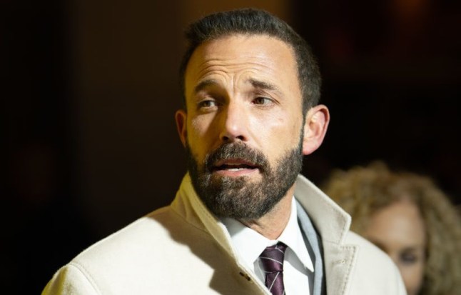 Ben Affleck in a beige jacket looks to the left while on stage.
