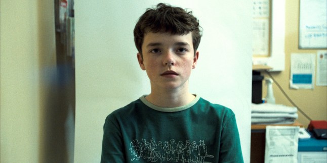 Owen Cooper as Jamie Miller in Adolescence having a photo taken at a police station. He's wearing a green T-shirt